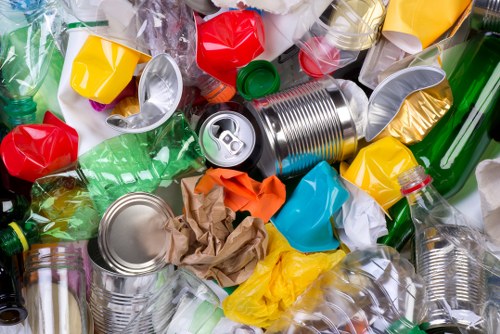 A densely packed collection of recyclable waste materials, including crushed aluminum cans, crumpled brown paper, and various plastic bottles in different colors such as green, red, yellow, and clear, all scattered on a surface likely outdoors or in a waste management environment. The aluminum cans are metallic with reflective surfaces, some partially dented, positioned centrally and leaning against surrounding plastic bottles which have different textures with smooth, semi-transparent finishes. The plastic bottles include some with caps intact, others without, and are of different shapes and sizes. The brown paper appears wrinkled and loosely layered, contrasting with the shiny plastic and metallic surfaces. The scene suggests an organized collection process typical of rubbish removal services like those provided by Commercial Waste Merton within the Merton postcode area, with a focus on waste segregation and disposal. Bright lighting highlights the varied colors and textures of the waste materials, emphasizing the volume and diversity of household or commercial rubbish intended for collection and recycling.