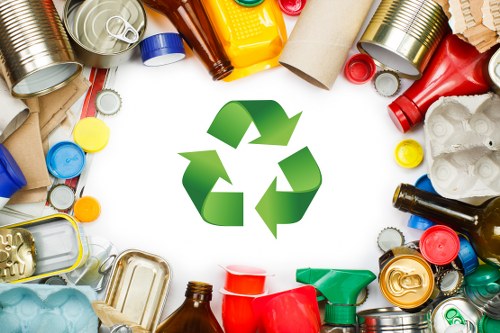 A collection of various discarded packaging materials and containers arranged in a circular pattern around a central green recycling symbol. The items include metal cans with silver, red, and yellow tops, plastic bottles in shades of clear, brown, and green, along with plastic lids, caps, and tubing. Some containers are crushed or flattened, revealing textures such as smooth, reflective metal surfaces and matte plastic finishes. Paper and cardboard tubes are visible among the waste, some partially rolled or torn. The waste materials are placed on a neutral, light-colored surface, likely outdoors or in a waste management setting, with the recycling symbol prominently centered to suggest proper disposal. The scene reflects typical rubbish associated with domestic or commercial waste, aligning with rubbish removal services provided by companies like Commercial Waste Merton, especially around urban areas like Merton. The natural lighting highlights the varied textures and colors, emphasizing the importance of recycling and waste management in reducing landfill contribution in the local area.