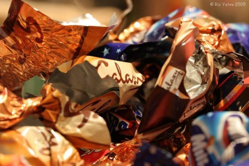 Close-up photograph of a pile of crumpled sweet and chocolate wrappers in various metallic, foil, and paper finishes, displaying colours such as copper, silver, blue, and brown. The wrappers are scattered and layered over each other, with some partially flattened and others more tightly crumpled, indicating recent disposal. The background is blurred, suggesting an indoor or outdoor environment, possibly on a pavement or table surface. The textures range from reflective and shiny to matte paper, with identifiable partial logos and branding visible on some wrappers, emphasizing waste materials related to confectionery packaging. The scene represents typical rubbish collected by Commercial Waste Merton during a general waste collection or rubbish removal service in Merton, contributing to environmental cleanliness and waste management efforts in the local area.