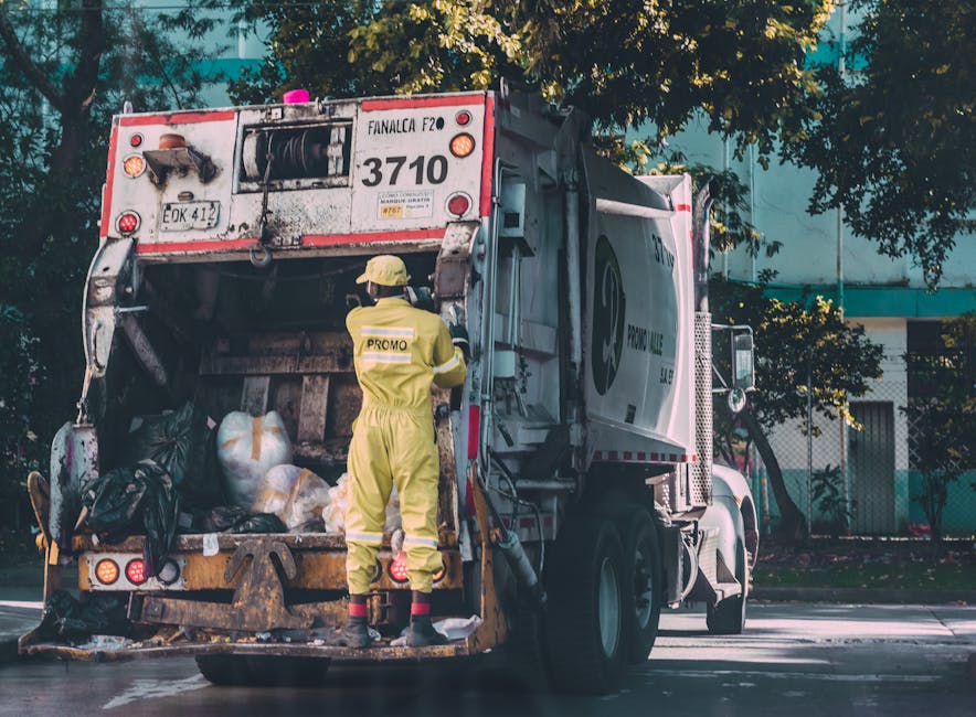 A waste collection truck, painted in white and red, is positioned on an urban street with its rear door open, revealing a collection of black and white trash bags packed inside. A worker wearing a yel