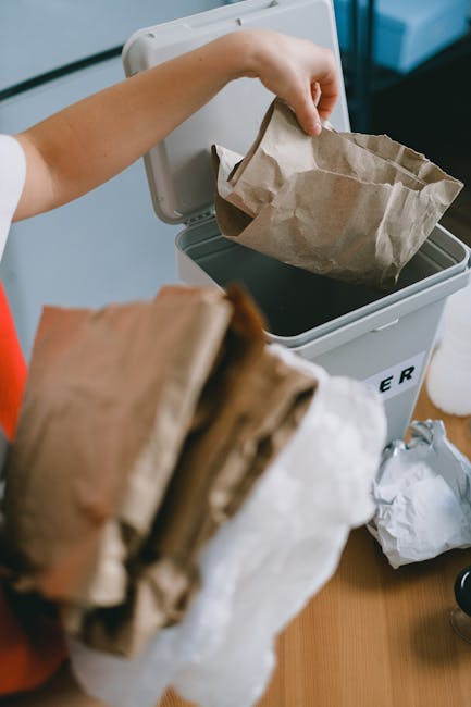 A waste collection truck, painted in white and red, is positioned on an urban street with its rear door open, revealing a collection of black and white trash bags packed inside. A worker wearing a yel
